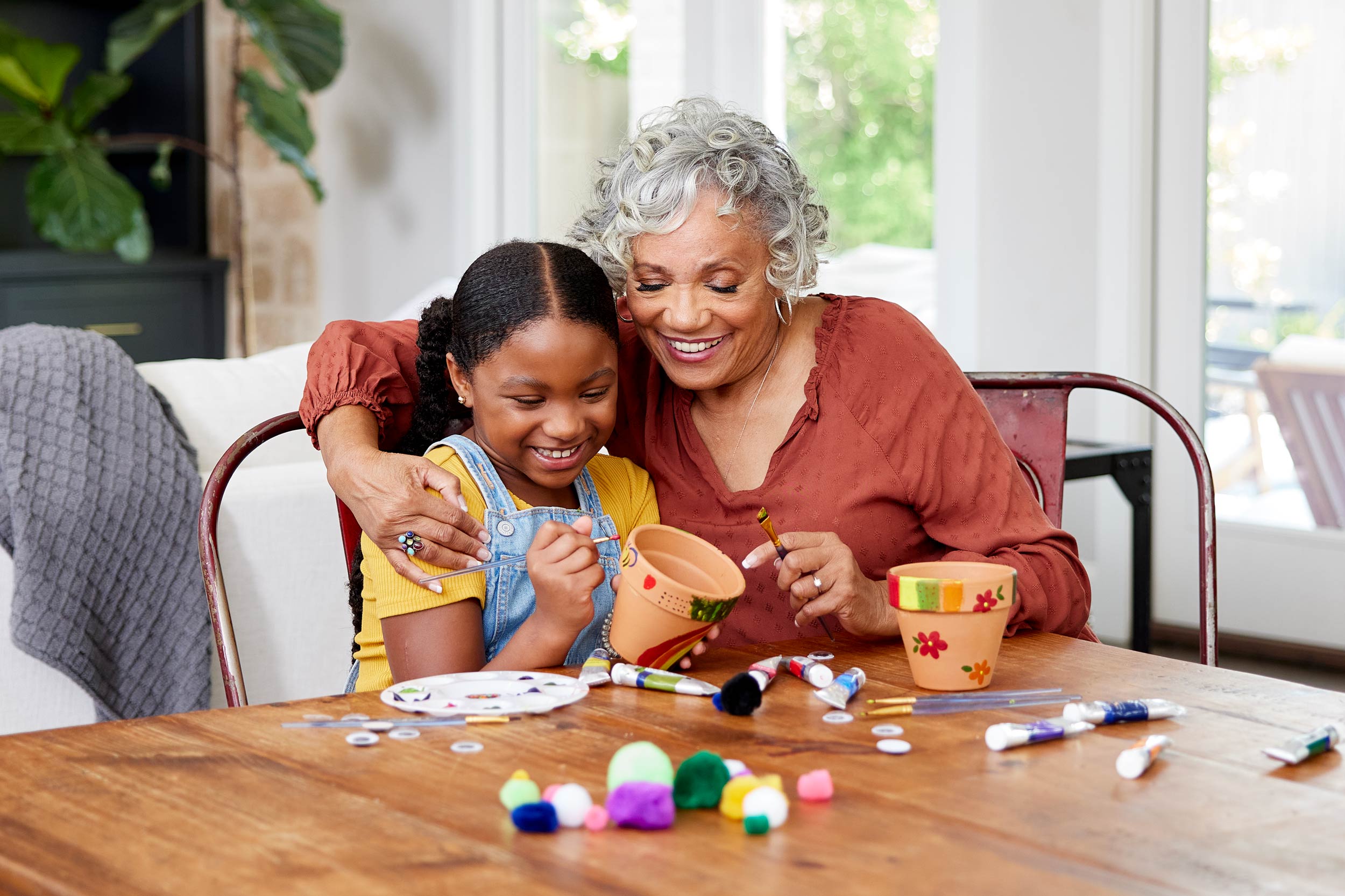 Indoor lifestyle photograph of grandmother and granddaughter crafting art together at dining room table.