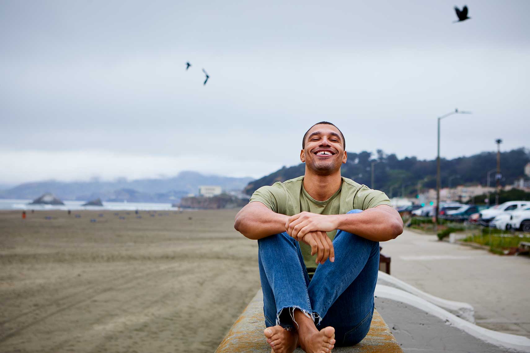 Outdoor lifestyle photograph of a young man sitting near the beach on a cloudy day with birds in the sky.